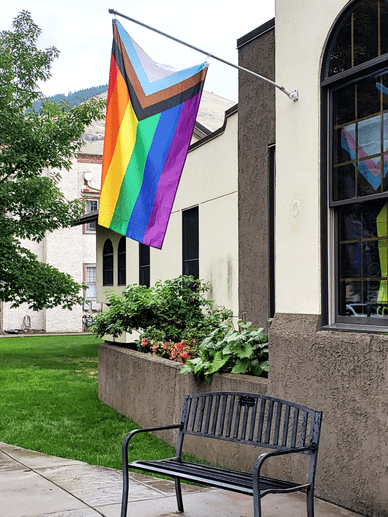 Progressive Pride flag hanging above a metal bench, in front of UCC Missoula